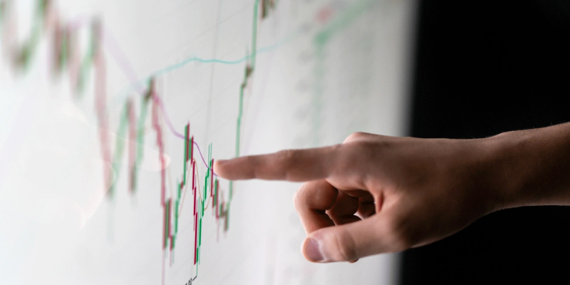 Hand pointing at a green and red line chart on a screen, dark background