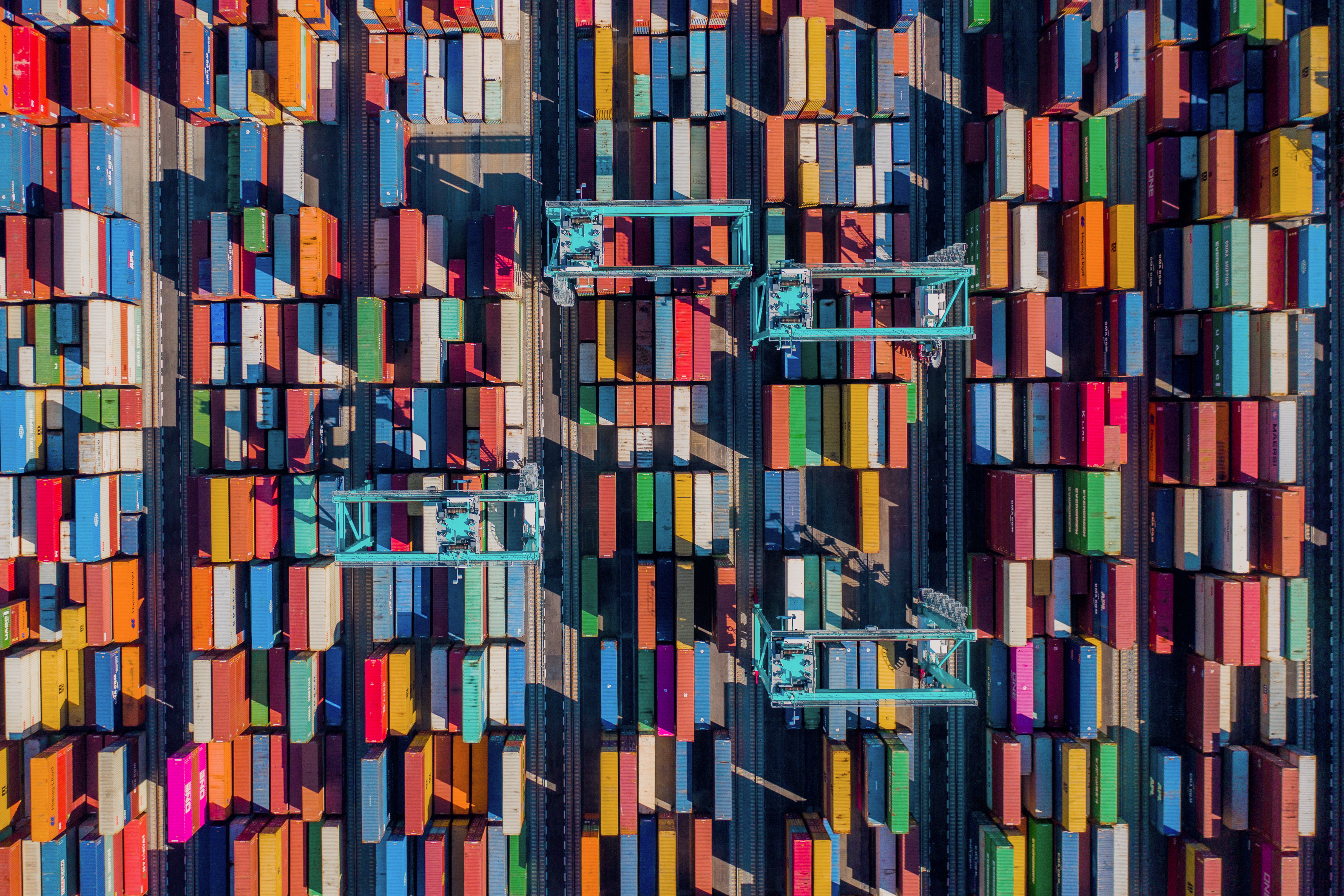 Aerial view of stacked shipping containers at a cargo port with cranes loading and unloading freight.