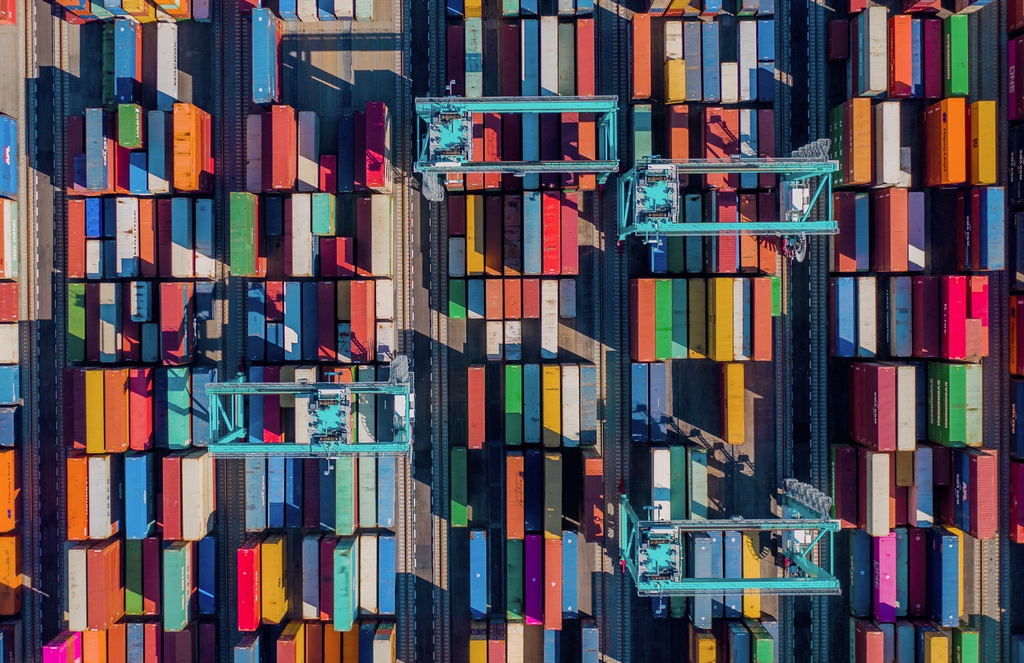 Aerial view of stacked shipping containers at a cargo port with cranes loading and unloading freight.
