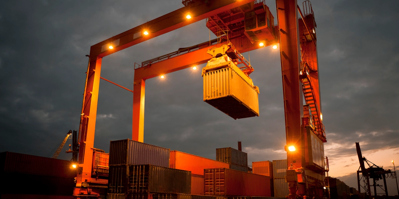 Gantry crane lifting a shipping container over stacked containers at a port at dusk