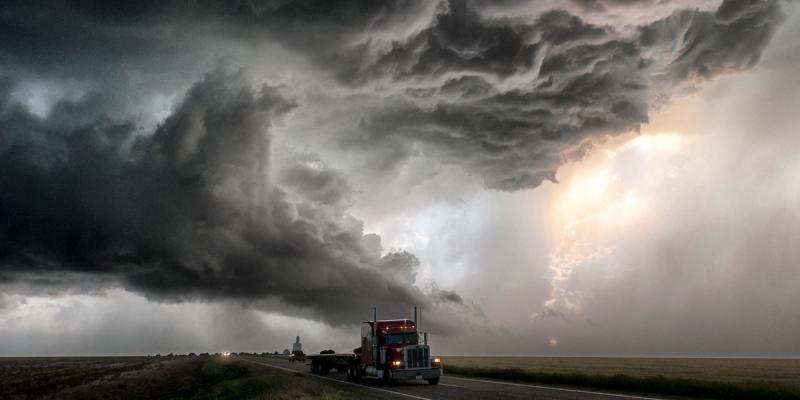 A red semi-truck with flatbed trailer drives a highway beneath a vast dark supercell storm in flat farmland.