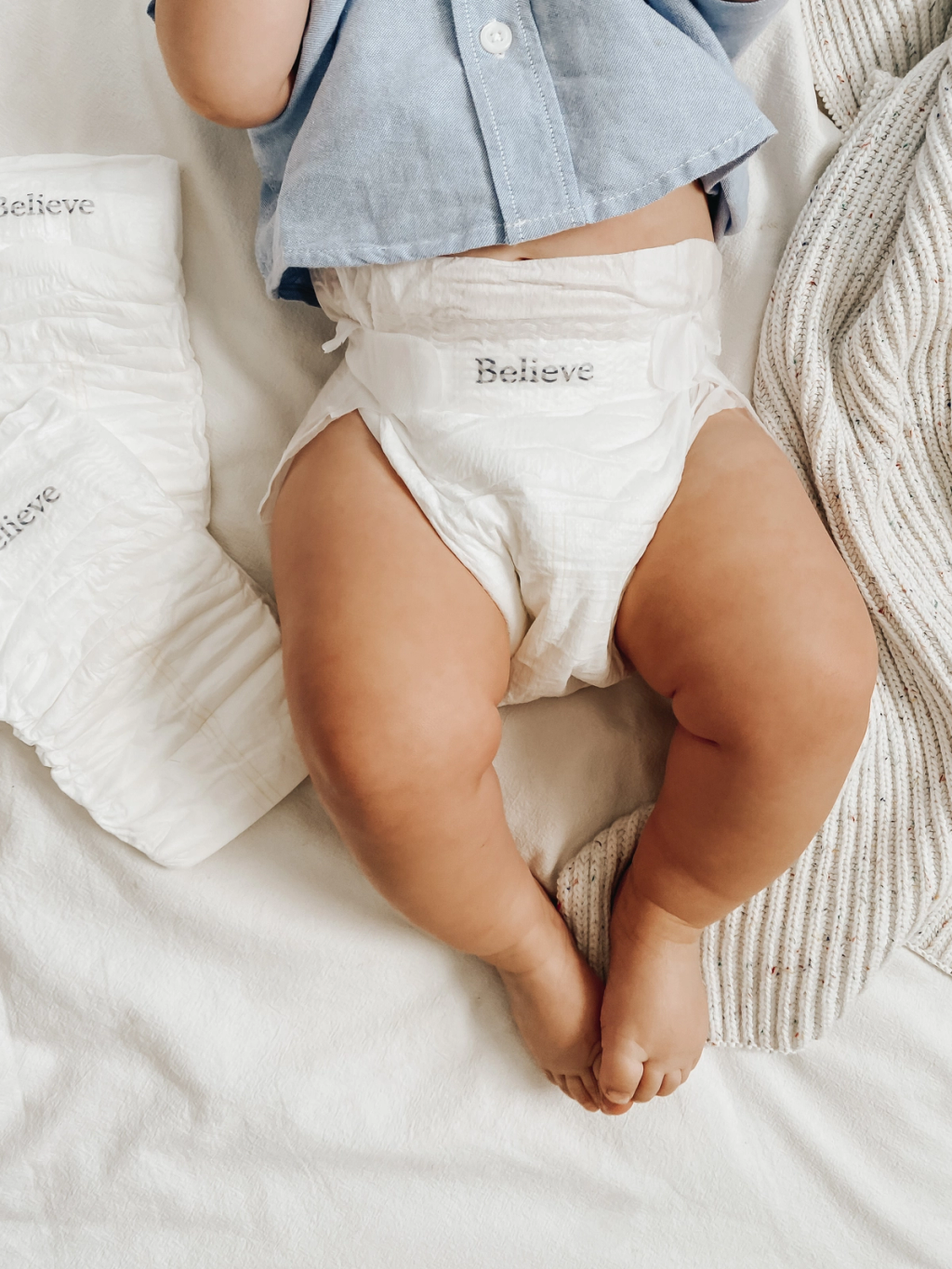 Baby lying on a bed wearing a Believe diaper, surrounded by extra diapers