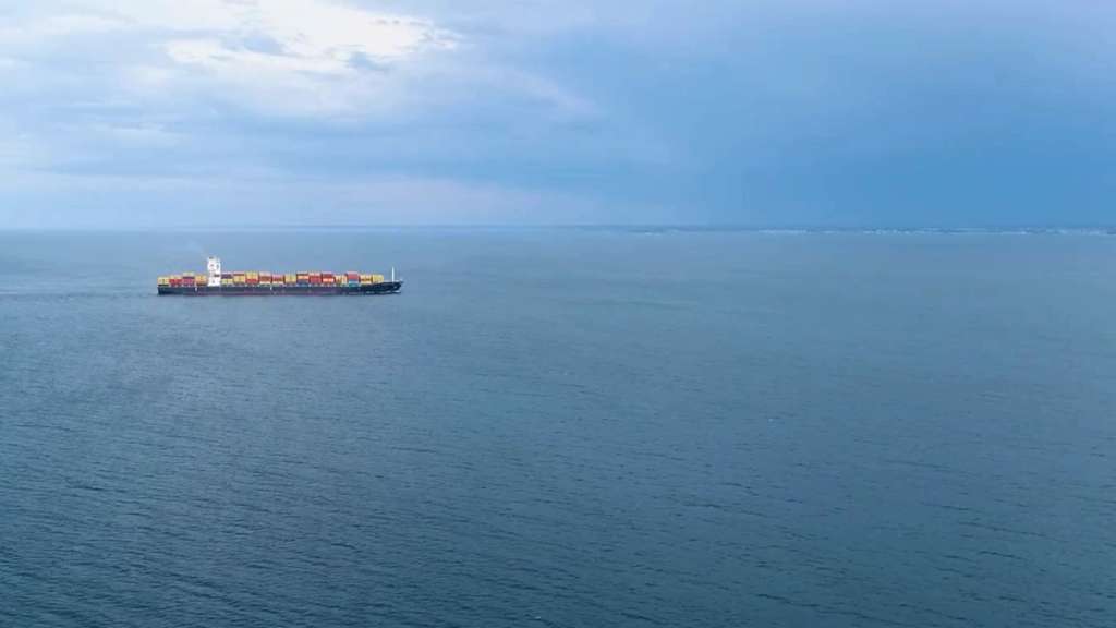 Aerial view of a container ship sailing across open ocean, leaving a wake trail behind it.