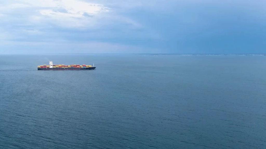 Aerial view of a container ship sailing across open ocean, leaving a wake trail behind it.