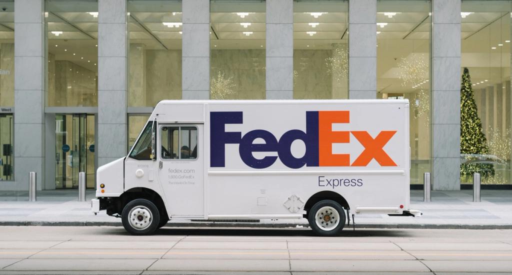 White FedEx Express delivery truck parked at a curb in front of a building with a lit holiday tree