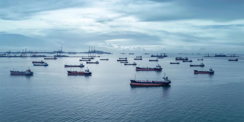 Aerial view of dozens of cargo and tanker ships anchored offshore near a port under a cloudy sky