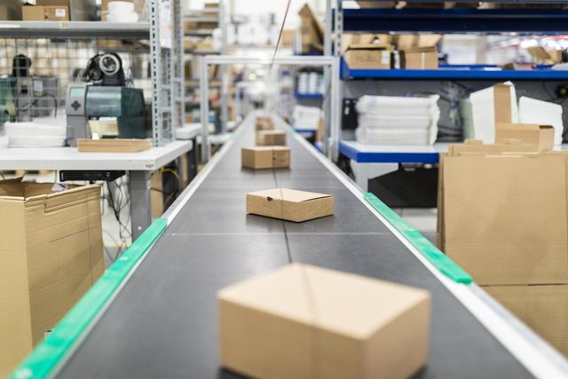 A small cardboard box travels along a black conveyor belt in a warehouse lined with shelved boxes.