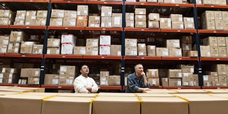 Two men in a warehouse standing among stacked cardboard cartons before tall shelves of labeled boxes