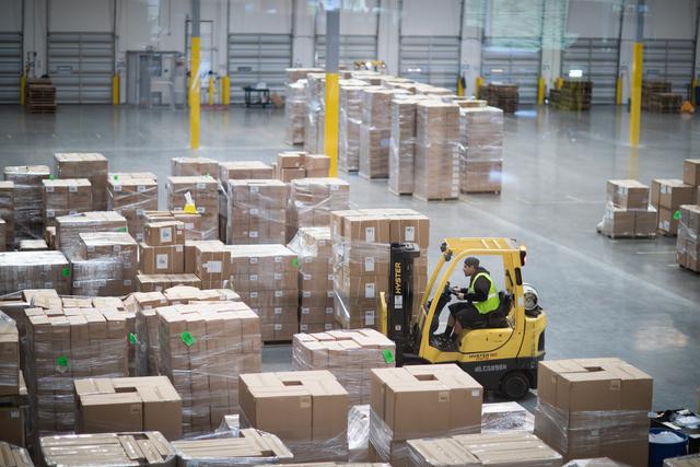 Worker drives a yellow Hyster forklift among rows of palletized cardboard boxes in a warehouse