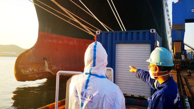 Two dock workers in hard hats and protective gear stand beside a container ship's hull at a port, one pointing ahead
