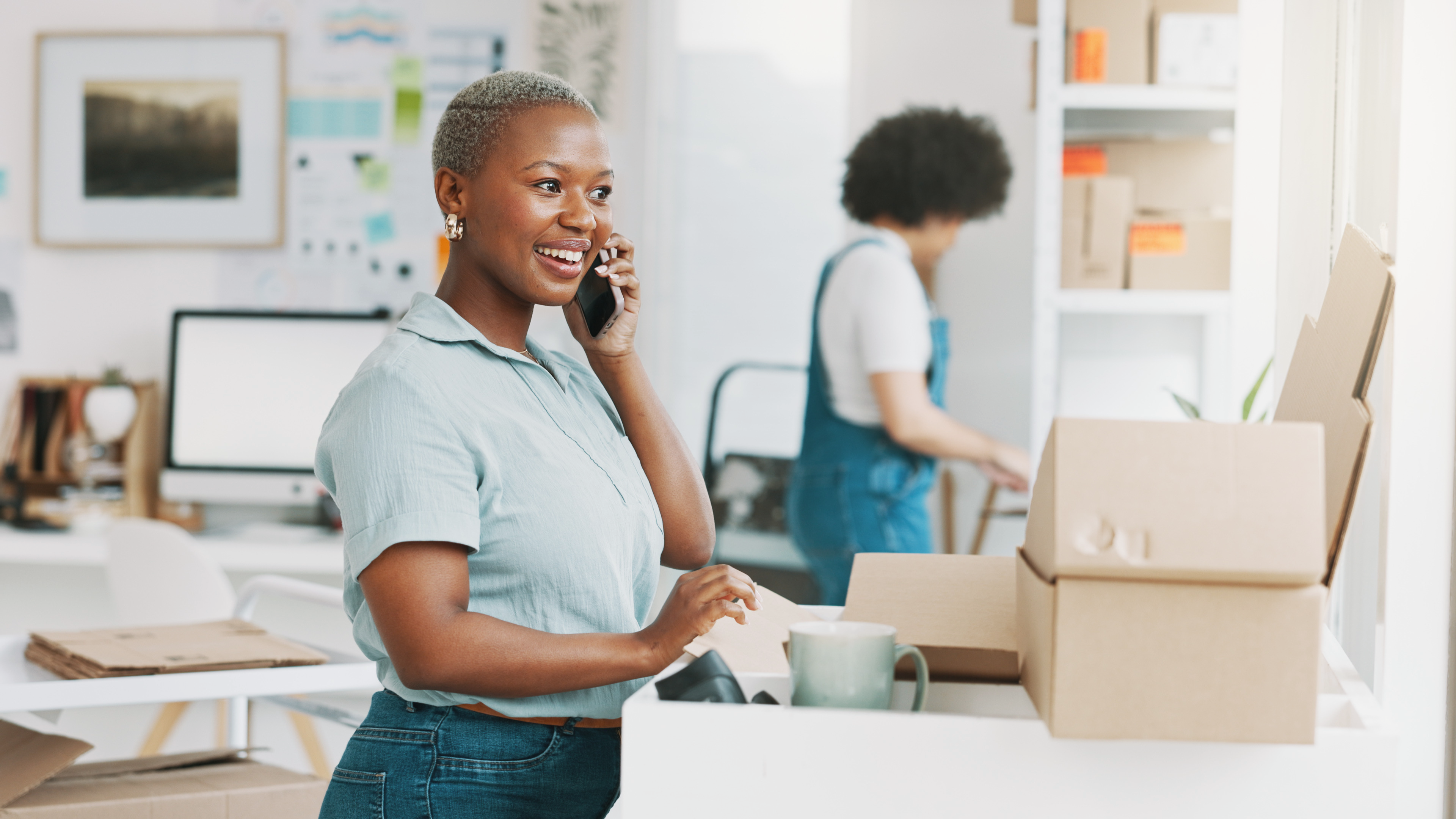 Woman in an office talking on a phone while holding a cardboard box during a move or delivery task.