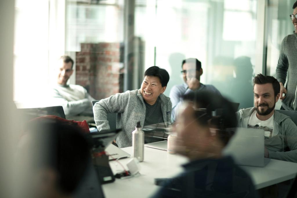 Smiling colleagues seated around a table with laptops in a bright office, viewed through glass