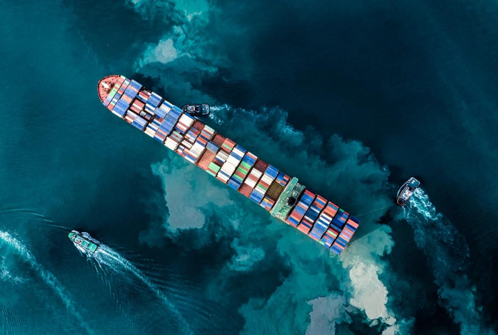Aerial overhead view of a fully loaded container ship escorted by tugboats on green-blue sea