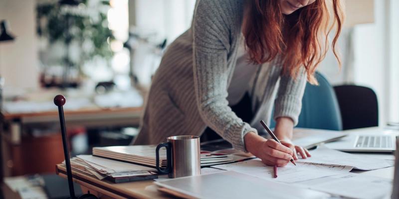 Woman with red hair leaning over a cluttered desk writing on documents next to a metal mug and laptop