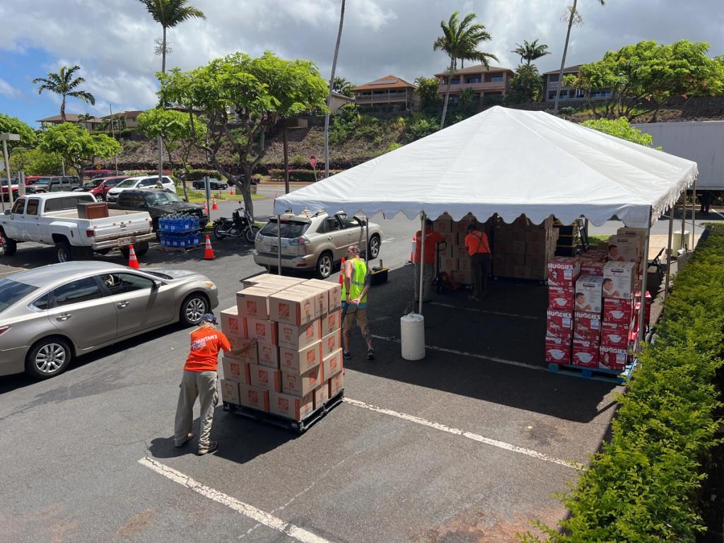 Volunteers in orange vests load boxed supplies onto a cart near a white relief tent in a Hawaii parking lot.