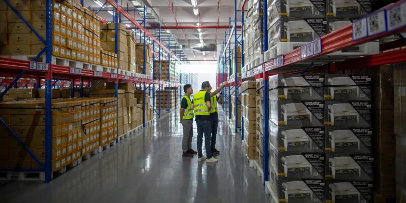 Three workers, one in a hi-vis vest, talk in the aisle of a warehouse with tall racks of pallets and boxes.