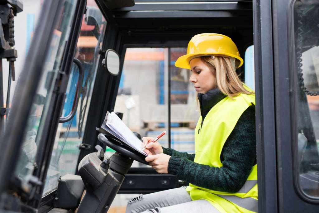 Heavy equipment operator in safety gear checks clipboard inside vehicle cab at construction site.