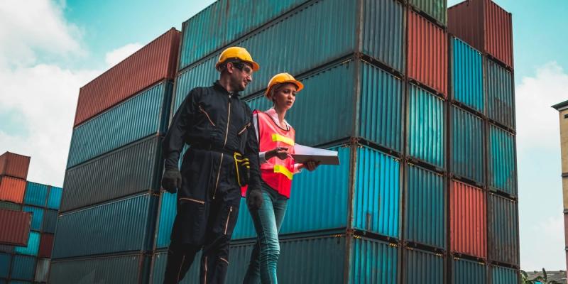 Two workers in hard hats and hi-vis vests reviewing a document in front of stacked shipping containers