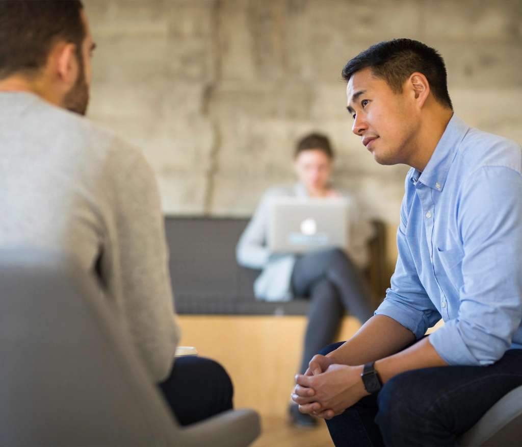Two colleagues talking in a relaxed office lounge while a third works on a laptop behind them