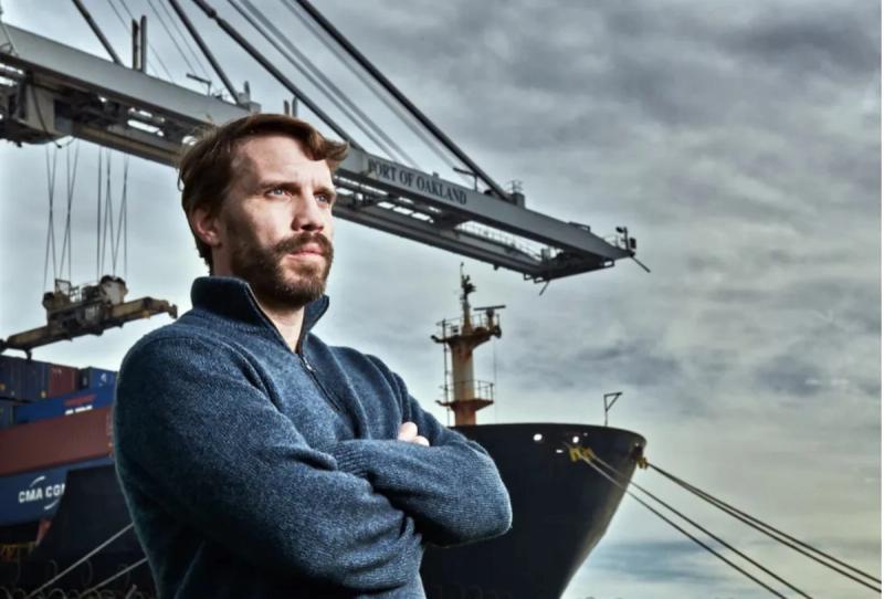 Bearded man with arms crossed stands before a Port of Oakland crane and the bow of a docked container ship.
