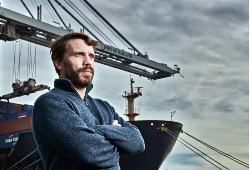 Bearded man with arms crossed stands before a Port of Oakland crane and the bow of a docked container ship.