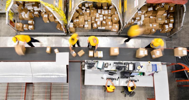 Overhead view of warehouse workers in yellow caps sorting and scanning packages along a conveyor and parcel chutes, motion-blurred