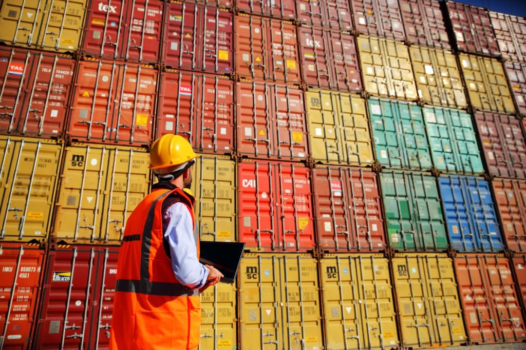 Worker in a hard hat and hi-vis vest holding a laptop before a towering wall of stacked shipping containers