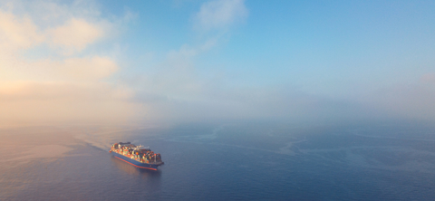 Aerial photo of a container ship sailing through low mist and fog over calm blue sea.