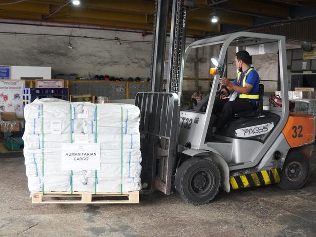 Forklift operator in a safety vest lifts a shrink-wrapped pallet labeled "Humanitarian Cargo" inside a warehouse.