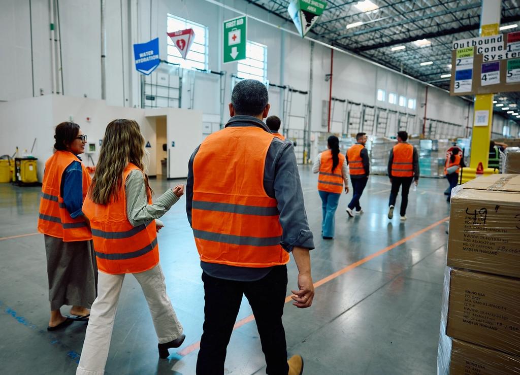 Workers wearing safety vests walk through a large industrial warehouse facility during a tour or inspection.