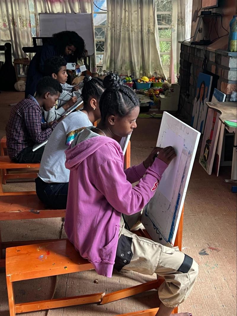 Young people seated on orange benches drawing and painting on large boards inside a community workshop