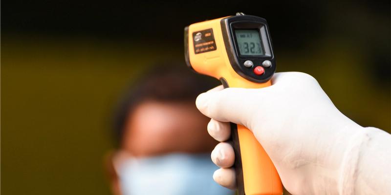 Gloved hand aiming an orange infrared thermometer reading 32 at a masked person in the background
