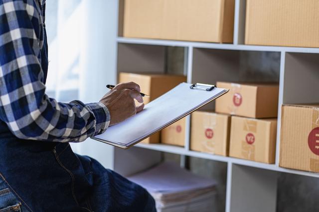 Worker in plaid shirt writing on a clipboard beside shelves of cardboard parcel boxes in a warehouse