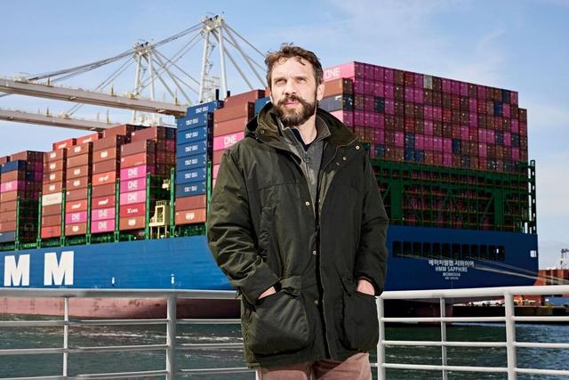 Bearded man in green jacket stands at a waterfront railing before the HMM Sapphire container ship and dockside cranes.
