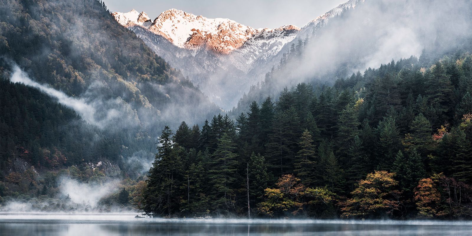 Misty forested mountain valley with snow-capped peaks reflected in a calm lake