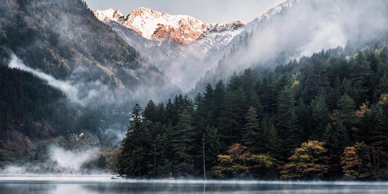 Misty forested mountain valley with snow-capped peaks reflected in a calm lake
