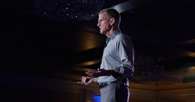 Man in a checked shirt speaking on a dimly lit conference stage with chandeliers overhead