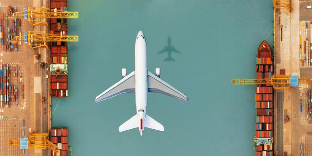 Aerial view of a white airplane flying over teal water between two container-stacked port terminals with cranes