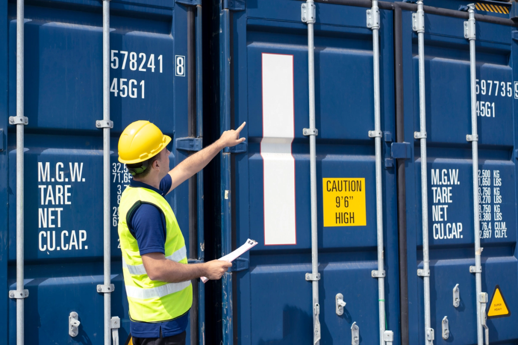 A port worker in a yellow hard hat and hi-vis vest holds a clipboard and points up at a row of blue shipping containers stencilled with tare and capacity markings.