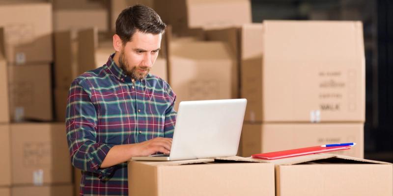 Bearded man in plaid shirt typing on a laptop set atop stacked cardboard boxes in a warehouse