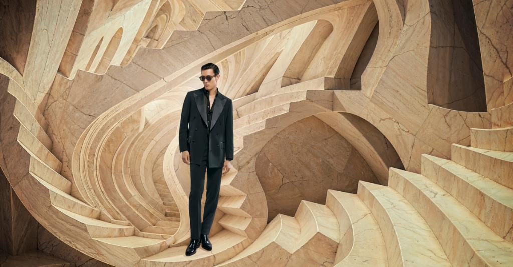 Man in a black tailored suit and sunglasses posing inside a sculptural sandstone-colored set