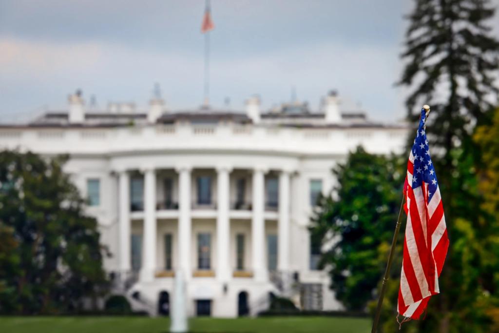 Tattered American flag in focus with the White House blurred behind it under a cloudy sky