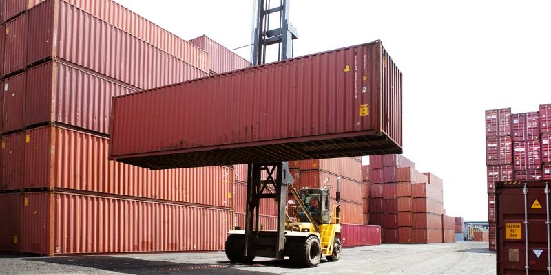 Yellow reach stacker lifting a maroon shipping container between rows of stacked containers
