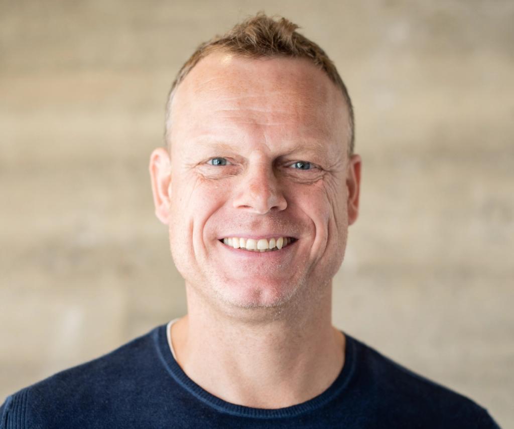 Headshot of a smiling man with short blond hair wearing a navy sweater against a concrete wall.