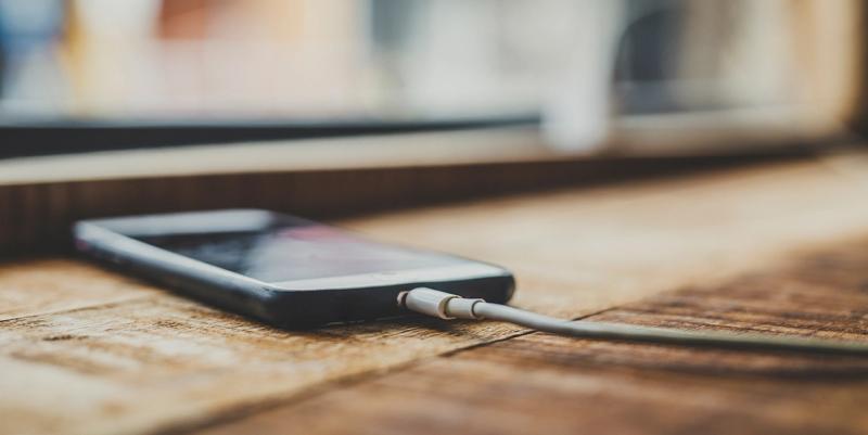 Smartphone lying face-up on a rustic wooden table with a white charging cable plugged in, blurred window behind.