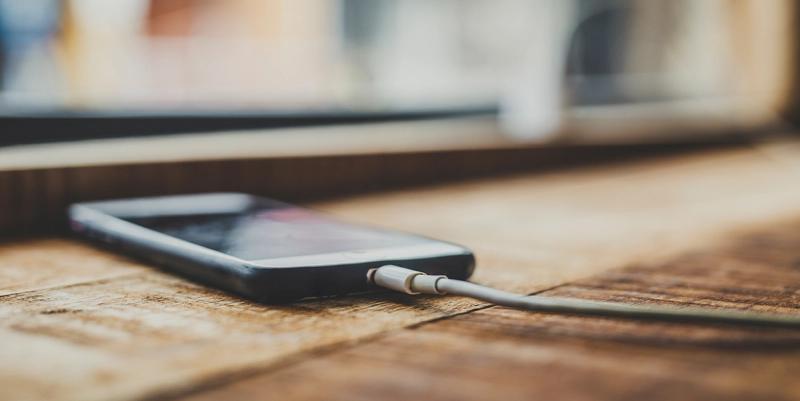 Smartphone lying face-up on a rustic wooden table with a white charging cable plugged in, blurred window behind.