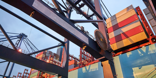 Crane spreader lifting a stacked shipping container at a busy port terminal at dusk, representing logistics scaling and funding
