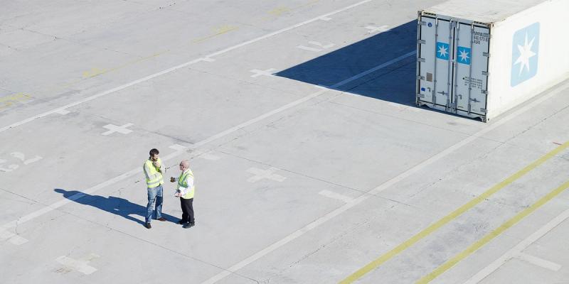 Two workers in hi-vis vests talk on a wide concrete yard beside a Maersk refrigerated container