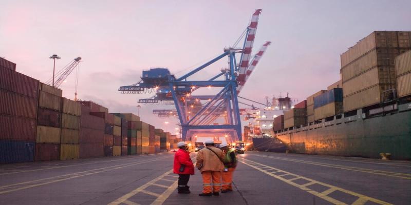 Three dockworkers in safety gear stand on a container terminal at dusk facing a ship and blue crane