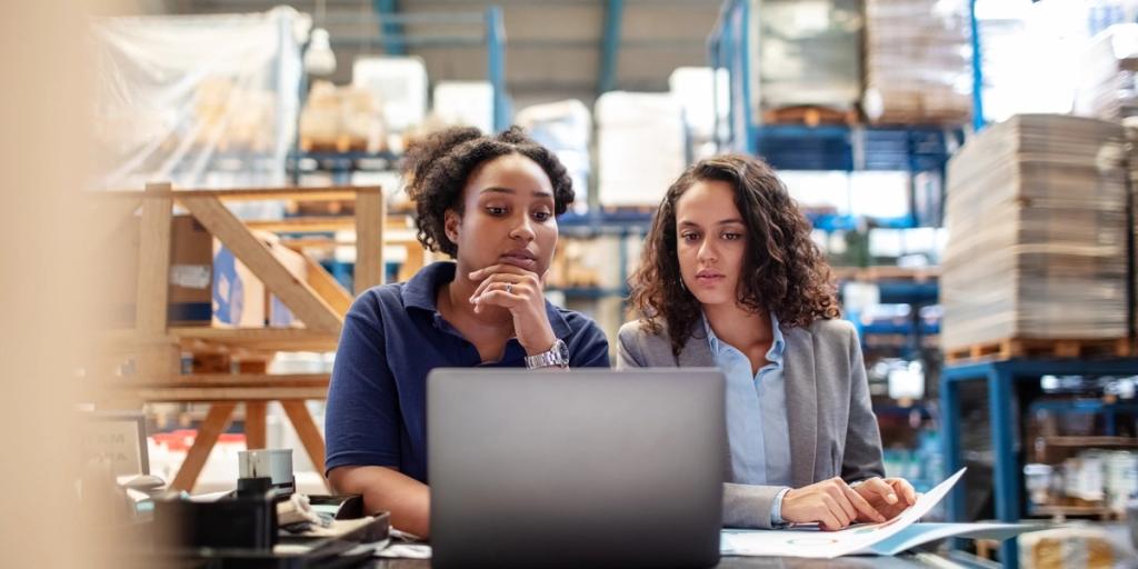 Two women study a laptop together at a warehouse desk surrounded by pallets and shelving